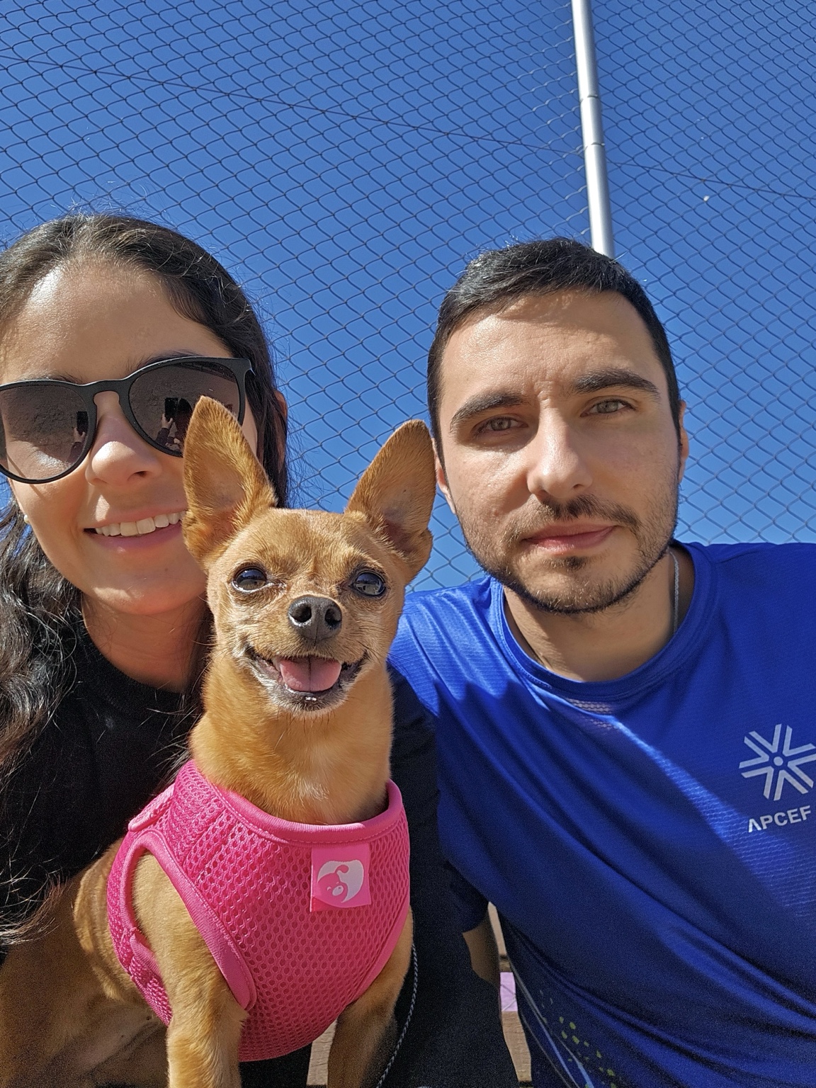 Selfie de Evelyn com Diego e a cachorrinha Mel, ao ar livre, em um dia de céu azul.