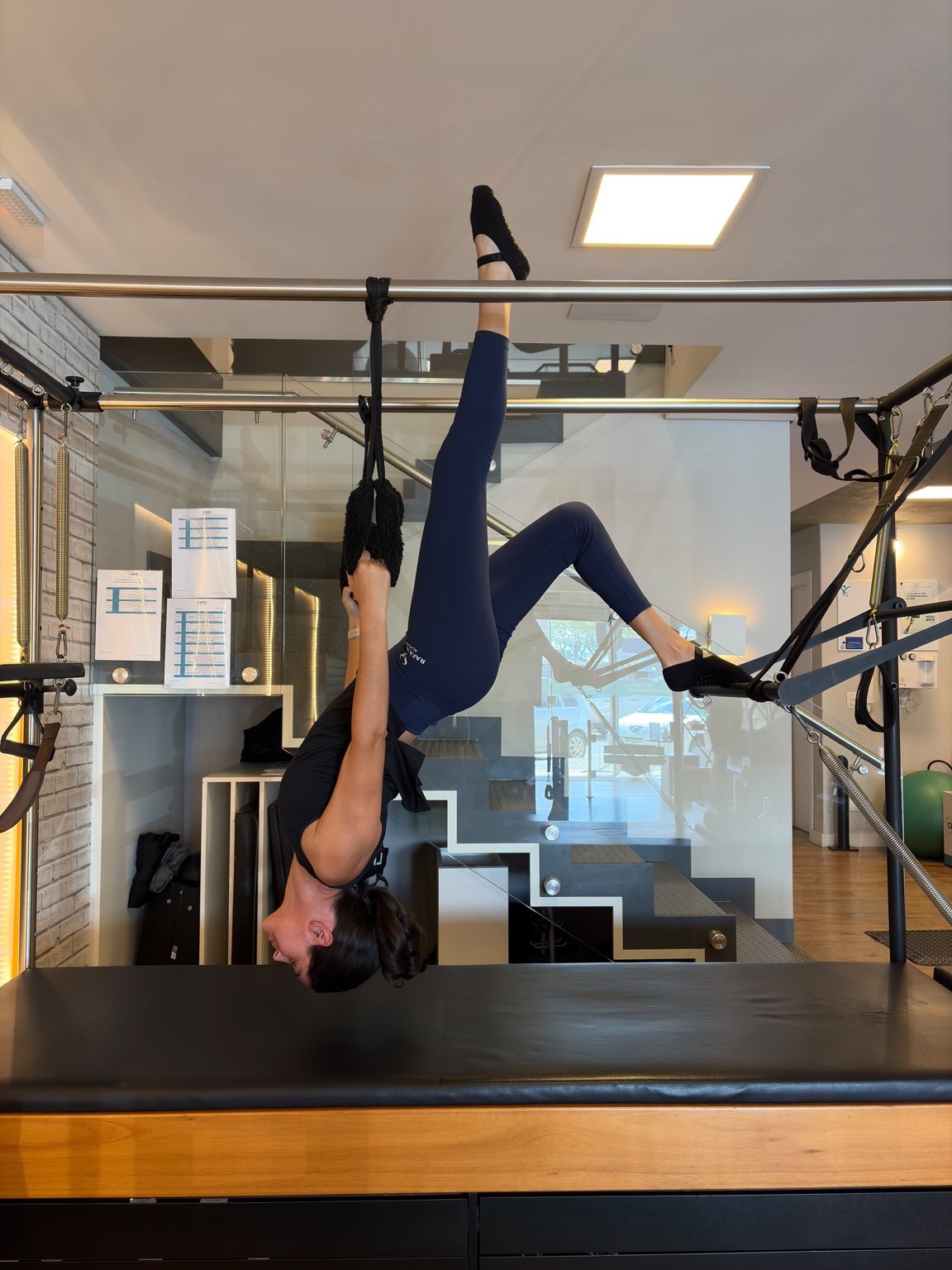 Evelyn praticando Pilates no aparelho cadillac, de cabeça para baixo, com uma perna estendida para cima.
